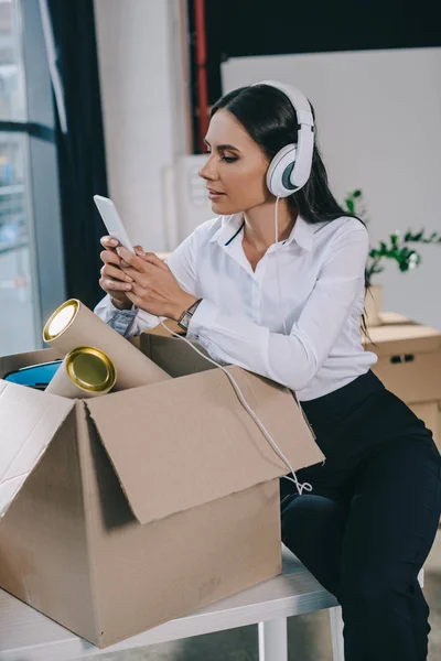 Beautiful young businesswoman in headphones using smartphone while unpacking box in new office — Stock Photo