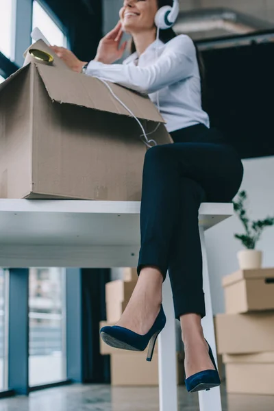 Low angle view of smiling young businesswoman in headphones using smartphone while unpacking box in new office — Stock Photo