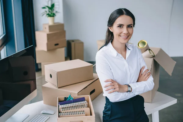 Joven mujer de negocios con los brazos cruzados sonriendo a la cámara mientras está sentada en la mesa en una oficina nueva - foto de stock