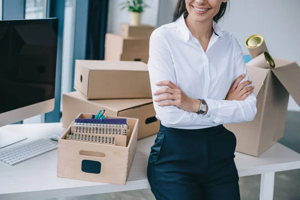 Обрезанный снимок улыбающейся деловой женщины со скрещенными руками, сидящей на столе в новом офисе — стоковое фото