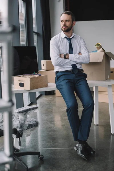 Hombre de negocios con los brazos cruzados sentado en la mesa y mirando hacia otro lado en la nueva oficina durante la reubicación - foto de stock