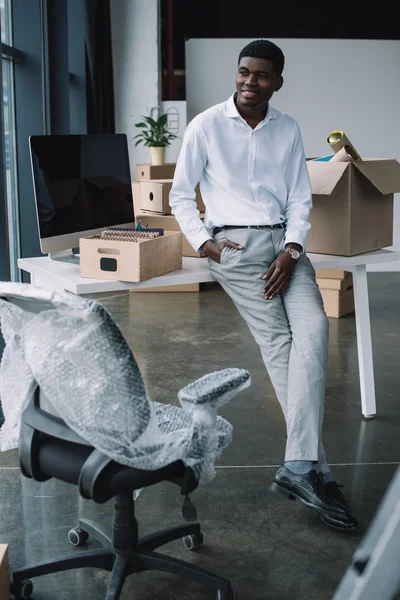 Sonriente joven hombre de negocios afroamericano sentado en la mesa con cajas y mirando hacia otro lado en la nueva oficina - foto de stock