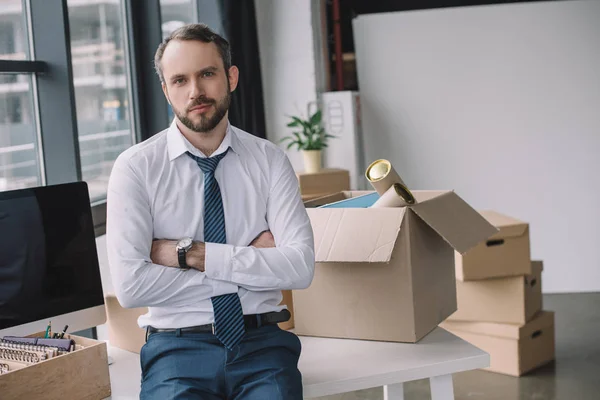 Geschäftsmann mit verschränkten Armen blickt in die Kamera, während er im neuen Büro auf einem Tisch mit Kisten sitzt — Stockfoto