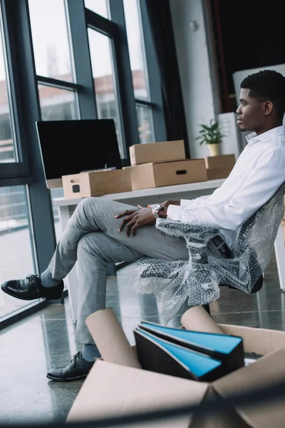 Joven hombre de negocios afroamericano mirando a la ventana mientras está sentado en una nueva oficina durante la reubicación - foto de stock