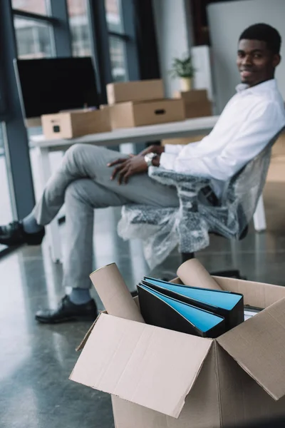 Vue rapprochée de la boîte en carton avec fournitures de bureau et homme d'affaires afro-américain assis derrière dans un nouveau bureau — Photo de stock