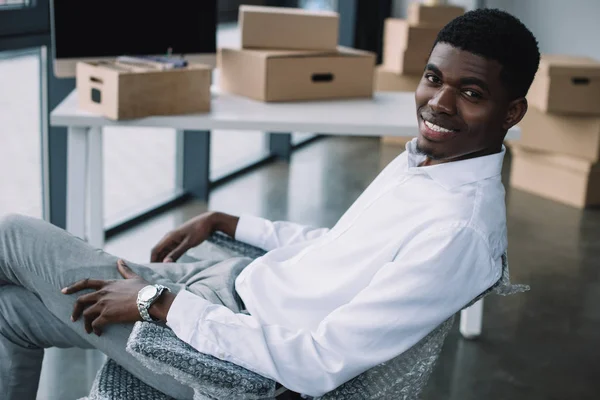 Heureux jeune homme d'affaires afro-américain souriant à la caméra tout en étant assis dans un nouveau bureau lors de la relocalisation — Photo de stock