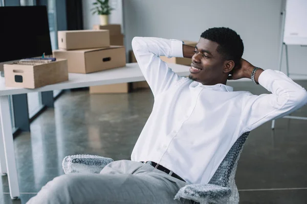 Smiling young african american businessman sitting with hands behind head and looking away in new office — Stock Photo