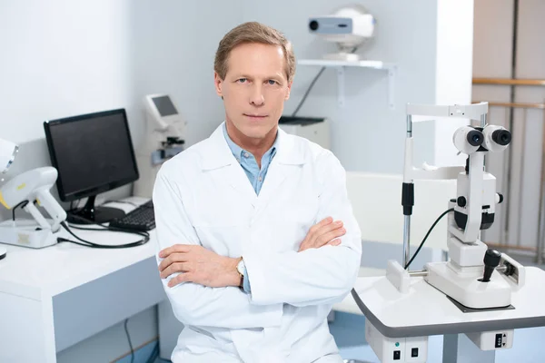 Augenarzt sitzt mit verschränkten Armen neben Spaltlampe im Sprechzimmer — Stockfoto