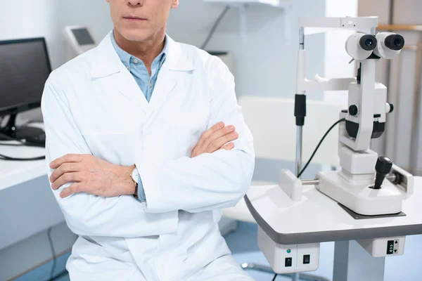 Cropped image of ophthalmologist sitting with crossed arms near slit lamp in consulting room — Stock Photo