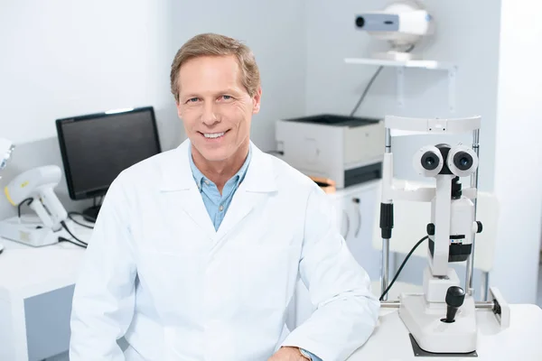 Guapo oftalmólogo sonriente mirando a la cámara en la sala de consulta - foto de stock