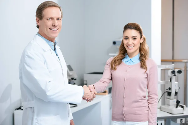Oftalmólogo sonriente y paciente estrechando las manos en la clínica - foto de stock