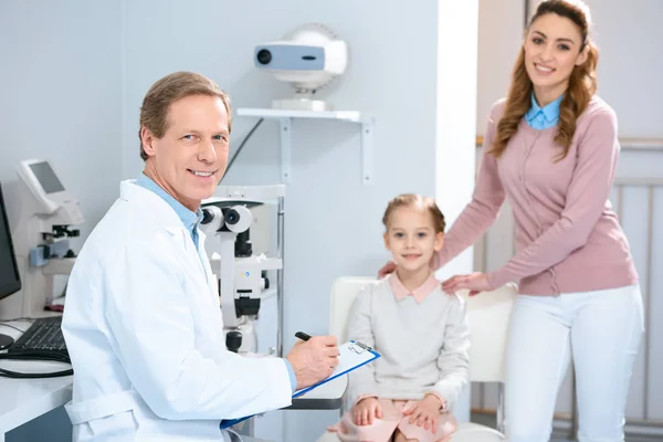 Sonriente madre, hija y oftalmólogo mirando a la cámara en la sala de consulta oculista - foto de stock