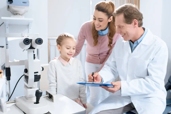 Lächelnder Augenarzt schreibt Patienten im Sprechzimmer etwas ans Klemmbrett — Stockfoto