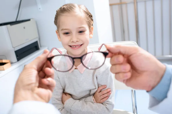 Обрезанный образ офтальмолога, предлагающего новые очки улыбающемуся ребенку в клинике — стоковое фото