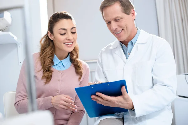 Beau médecin montrant quelque chose dans le presse-papiers au patient souriant à la clinique — Photo de stock