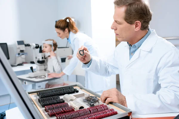 Opticien tenant des lentilles pour cadre d'essai tout en collègue examinant enfant avec test de champ visuel — Photo de stock