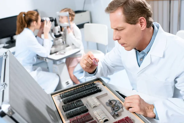 Masculino oculista segurando lentes para julgamento quadro enquanto colega examinando criança com teste de campo visual — Fotografia de Stock