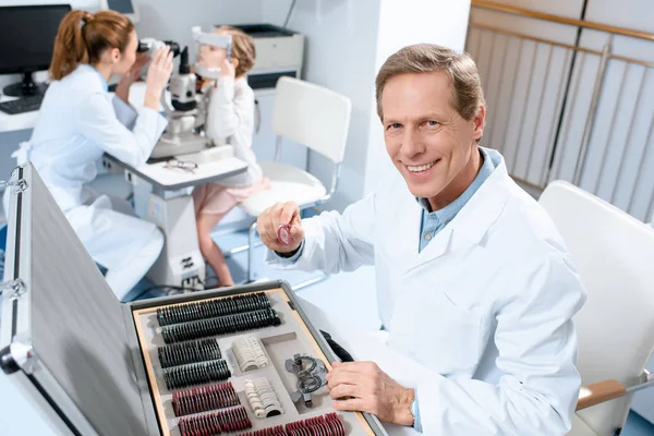 Male optometrist holding lenses for trial frame while colleague examining kid with visual field test — Stock Photo