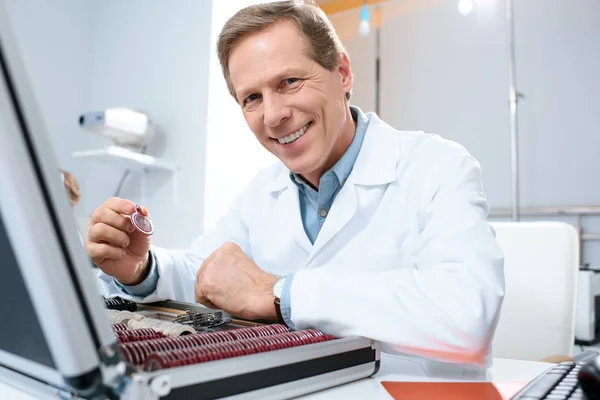 Óptica masculina sonriente sosteniendo lentes para el marco de prueba en la clínica - foto de stock