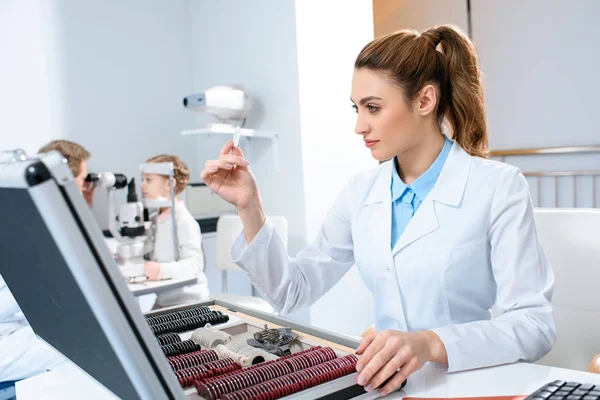 Female ophthalmologist with lenses for trial frame while colleague examining kid with visual field test — Stock Photo