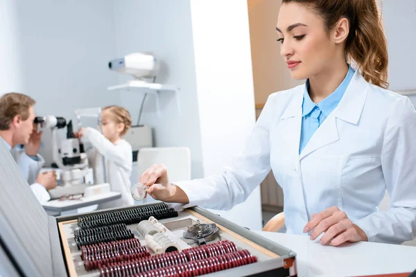 Female optometrist with lenses for trial frame while colleague examining kid with visual field test — Stock Photo