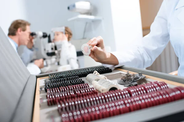 Vista recortada del oftalmólogo que sostiene las lentes para la montura del ensayo mientras su colega examina al niño con una prueba de campo visual - foto de stock