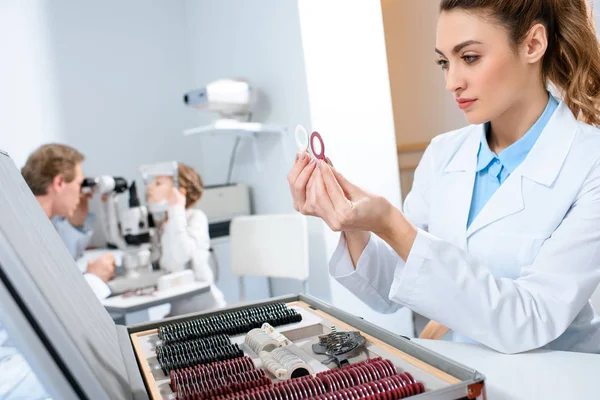 Female optometrist holding lenses for trial frame while colleague examining kid with visual field test — Stock Photo