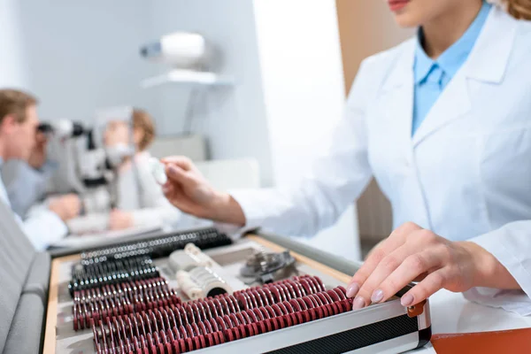 Cropped view of optician holding lenses for trial frame while colleague examining kid with visual field test — Stock Photo