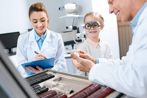 Oculists examining kid eyes with trial frame and writing diagnosis — Stock Photo