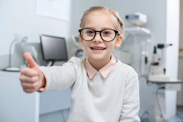 Smiling little child in eyeglasses showing thumb up in optical clinic — Stock Photo