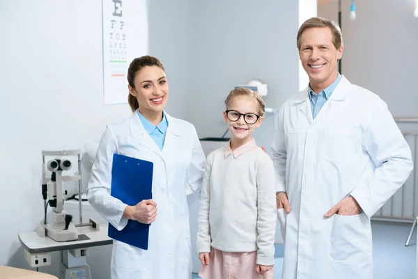 Two smiling ophthalmologists and little child in glasses standing in clinic — Stock Photo