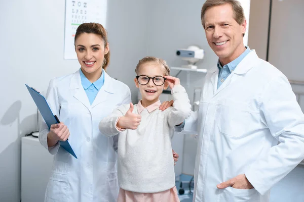 Dos optometristas sonrientes y un niño pequeño en gafas que muestran el pulgar hacia arriba - foto de stock