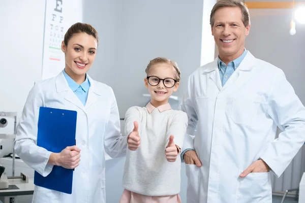 Optometristas sonrientes y un niño pequeño en gafas que muestran los pulgares hacia arriba - foto de stock