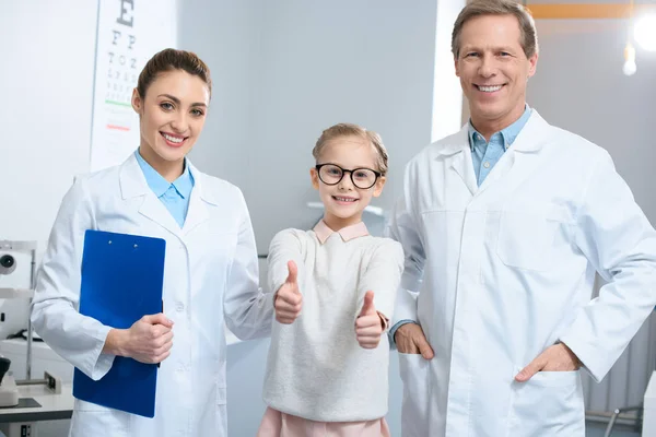 Optometristas sorridentes e criança pequena em óculos mostrando polegares para cima — Fotografia de Stock