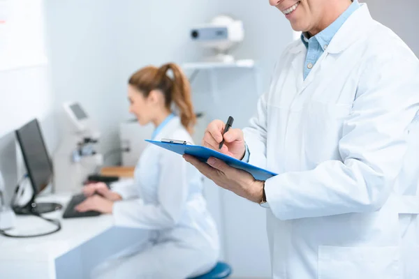 Cropped view of male optometrist writing diagnosis while colleague using with computer in clinic — Stock Photo
