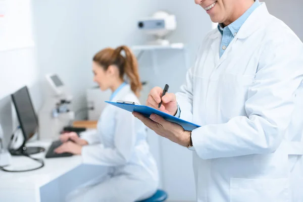 Zugeschnittene Ansicht eines männlichen Optometristen, der Diagnose schreibt, während ein Kollege mit dem Computer in der Klinik arbeitet — Stock Photo