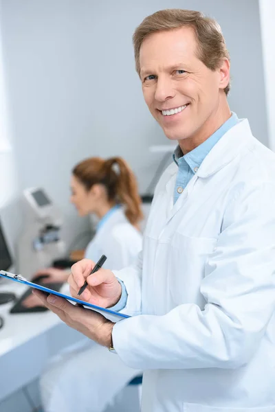 Opticien masculin écrivant le diagnostic tout en collaborant utilisant avec l'ordinateur dans la clinique — Photo de stock