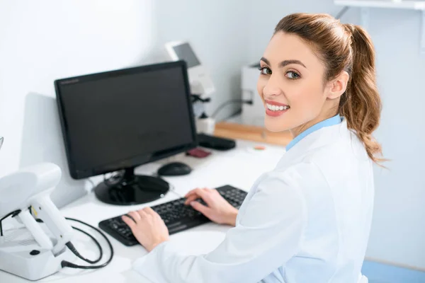 Lächelnd schöne Augenärztin, die mit dem Computer in der Klinik arbeitet — Stockfoto