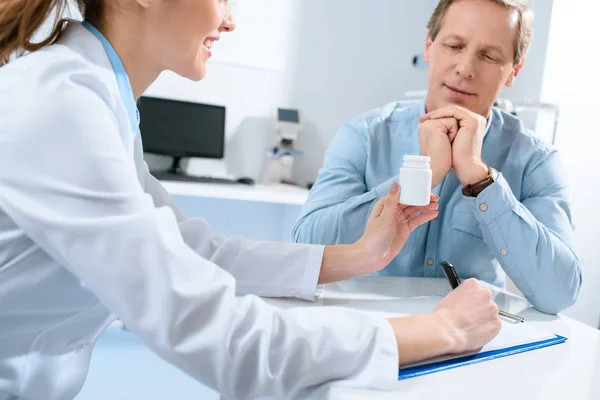 Médico sosteniendo botella de pastillas, escribiendo diagnóstico y hablando con el paciente maduro en la clínica - foto de stock