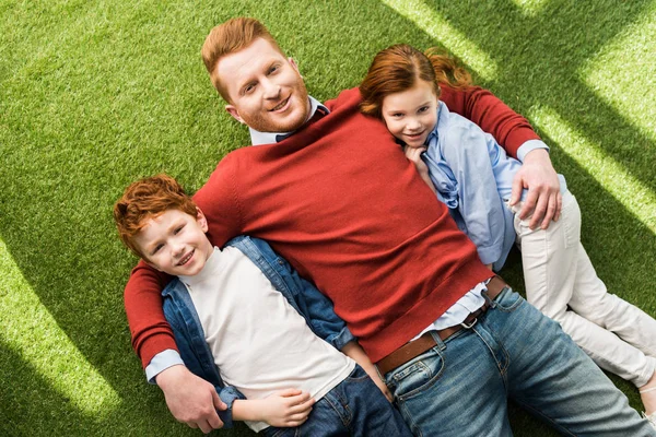 Ansicht von oben: glücklicher Vater mit entzückenden kleinen Kindern, die zusammen im Gras liegen und in die Kamera lächeln — Stockfoto