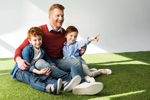 Heureux père avec mignons petits enfants assis ensemble sur l'herbe et regardant loin sur gris — Photo de stock