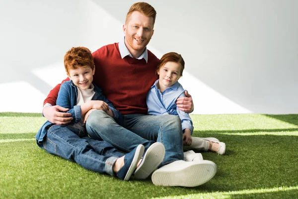 Feliz pelirroja padre y niños sentado juntos en la hierba y sonriendo a la cámara en gris - foto de stock