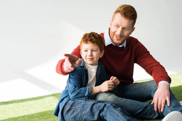 Feliz pelirroja padre e hijo sentado juntos y mirando hacia otro lado en gris - foto de stock