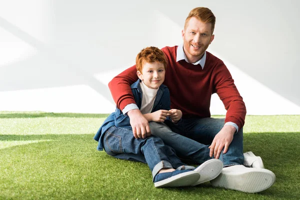 Happy father and son sitting on grass and smiling at camera on grey — Stock Photo