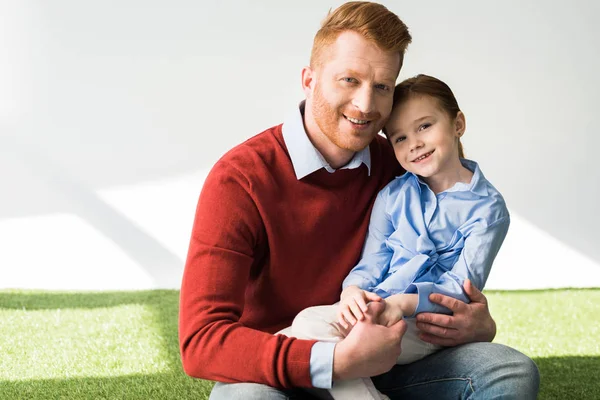 Happy redhead father and daughter sitting on grass and smiling at camera on grey — Stock Photo