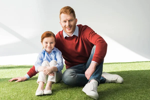 Glückliche rothaarige Vater und Tochter sitzen auf Gras und lächeln in die Kamera auf grau — Stockfoto