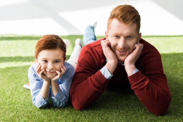 Happy redhead father and daughter lying on grass and smiling at camera on grey — Stock Photo