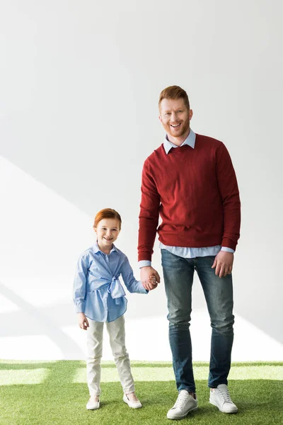 Happy redhead father and daughter holding hands and smiling at camera on grey — Stock Photo