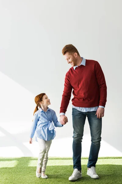 Счастливые рыжие отец и дочь держатся за руки и улыбаются друг другу на серой — стоковое фото