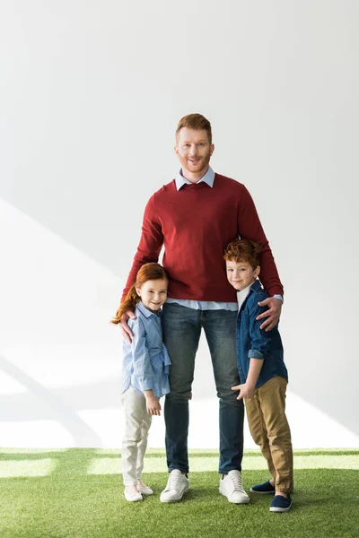 Feliz padre con adorable pelirroja niños de pie juntos en la hierba y sonriendo a la cámara en gris - foto de stock